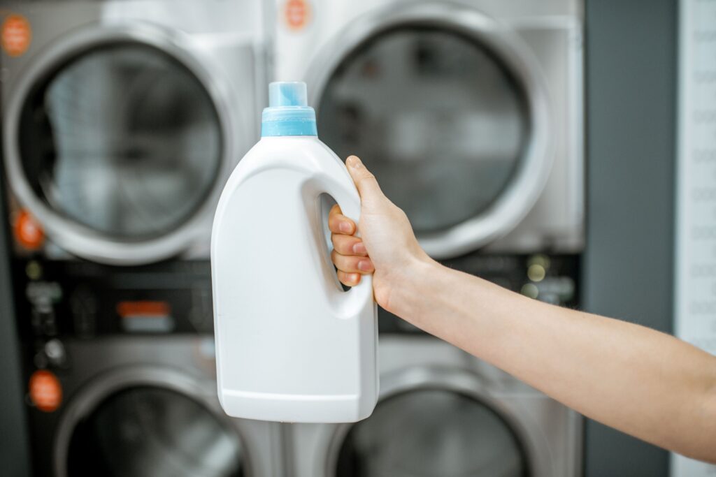 Hand mit Waschmittelflasche vor Waschmaschinen im SB-Waschsalon Nürnberg – Waschmittel ist inklusive